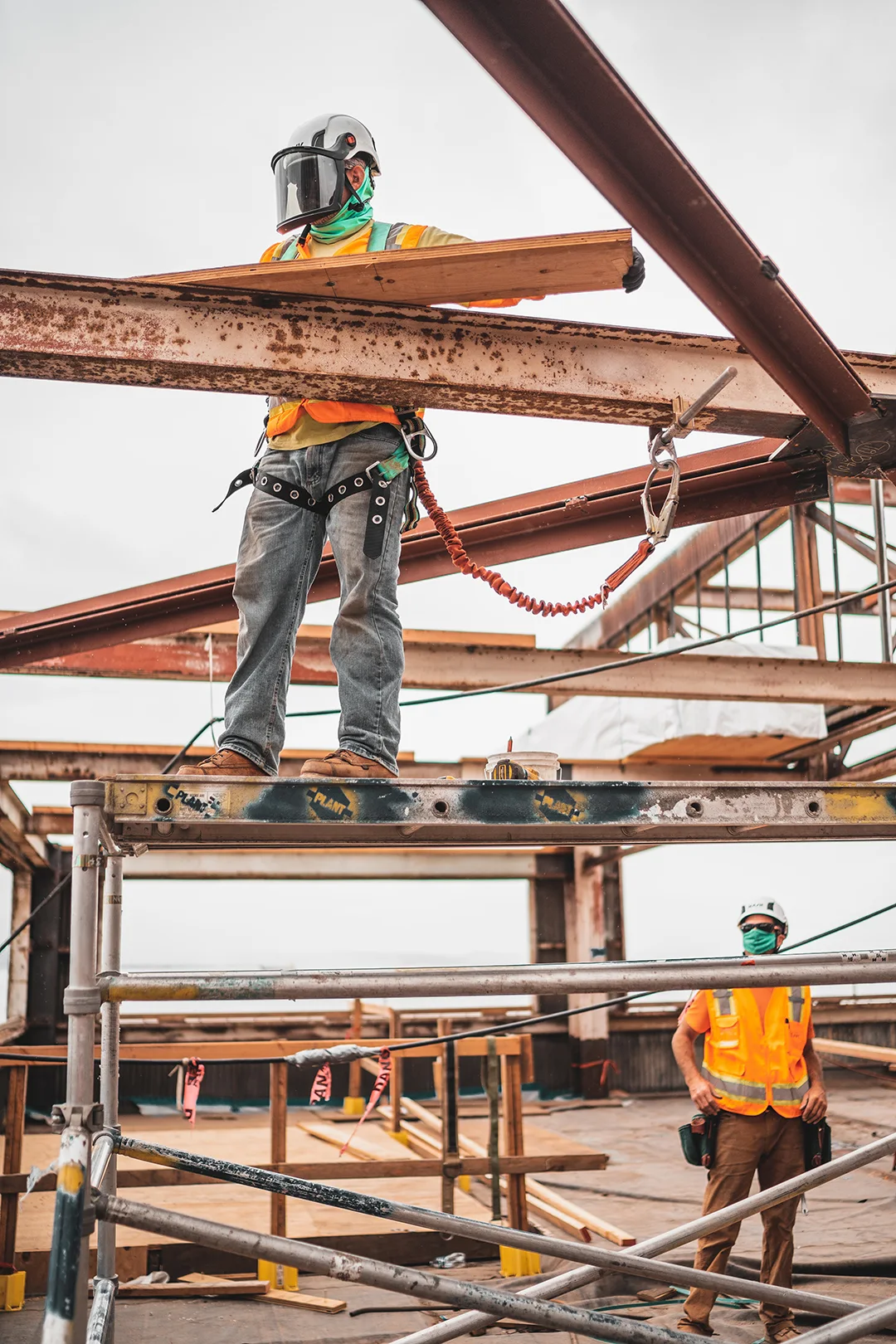 republix-commercial-products Construction workers wearing safety gear and helmets working on a steel structure, with one worker on a scaffold holding a wooden plank and secured with a safety harness.